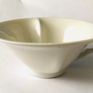 May include: A white ceramic teacup with a wide, shallow bowl and a single handle. The cup has a smooth, glossy finish.