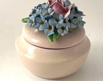 Puede incluir: Pequeña caja de cerámica rosa pálido con tapa. La tapa está adornada con un grupo de flores azules, una rosa rosa y hojas verdes. La caja tiene una forma suave y redondeada.