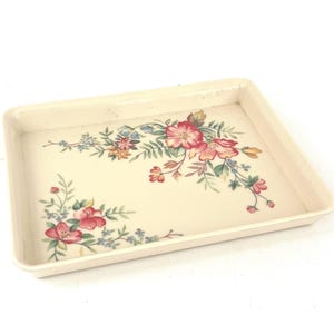 Pode incluir: Bandeja retangular creme com um desenho floral. A bandeja tem uma borda e é decorada com flores rosa, vermelhas, azuis e amarelas e folhas verdes. O padrão floral é disposto nos cantos.
