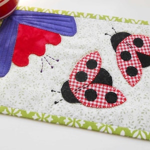 May include: A green and white fabric coaster with two red and black ladybugs with white polka dots. The coaster has a purple and red flower in the corner.