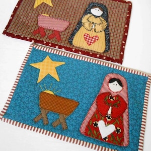 May include: Two handmade quilted Christmas ornaments featuring a nativity scene. The first ornament is brown and red with a star, manger, and a woman in a red robe. The second ornament is blue and red with a star, manger, and a woman in a red robe.
