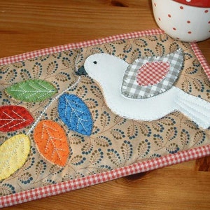 May include: A white fabric bird with a red and white checkered wing, perched on a branch with colorful leaves. The branch is on a brown fabric background with a red and white checkered border.