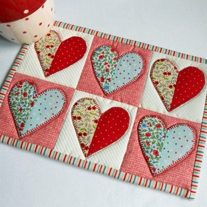 May include: A red and white quilted heart-shaped hot pad with a decorative border. The hot pad is made of fabric with a patchwork design and features six hearts in various colors and patterns.