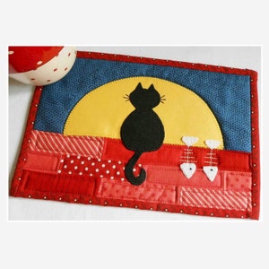 Op de afbeelding: Een rode, witte en blauwe patchwork stoffen onderzetter met een zwart katten silhouet dat op een gele maan zit. Twee witte vissen bevinden zich aan de onderkant van de onderzetter.