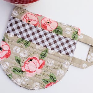May include: A quilted mug rug with a floral and gingham pattern. The mug rug features pink roses, green leaves, and a brown and white checkered section. The handle is a solid tan color with white floral accents.