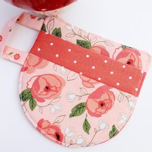 May include: A floral print pot holder with a handle. The pot holder features a light pink background with large pink roses and green leaves. A band of coral fabric with white polka dots is sewn across the top. The handle is light pink with small floral accents.