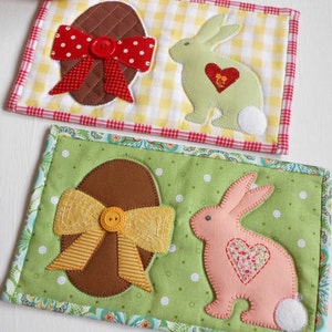 May include: Two handmade fabric coasters featuring a brown egg with a yellow bow and a pink bunny with a heart on a green background, and a brown egg with a red bow and a green bunny with a red heart on a yellow and red checkered background.