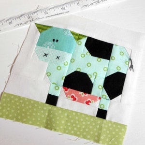 May include: A patchwork quilt block featuring a green, black, and pink cow with white polka dots. The cow is standing on a green and white polka dot background.