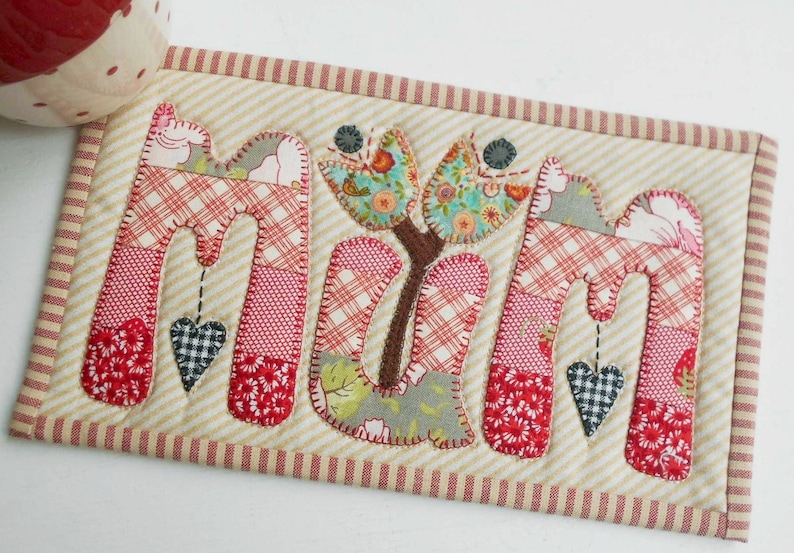 May include: A handmade fabric coaster with the word "MUM" spelled out in colorful patchwork letters. Each letter has a small heart hanging from it. A small floral design is in the center of the coaster.