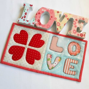 May include: A quilted fabric piece with a red and white striped border. The piece features a four-leaf clover design and the word "LOVE" spelled out with fabric letters. The letters and clover are in various patterns and colors.