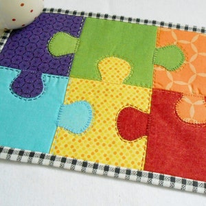 May include: A colorful puzzle-shaped coaster with a black and white checkered border. The coaster is made of fabric and has a patchwork design with different colors and patterns.