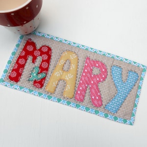 May include: A handmade fabric coaster with the name "MARY" spelled out in colorful patchwork letters. The letters are sewn onto a beige fabric background with a blue and white border.