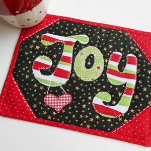May include: A red and black quilted coaster with a white, red, and green "Joy" design and a red and white gingham heart. The background is black with gold stars.