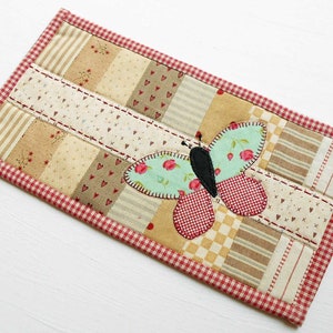 May include: A handmade quilted coaster with a patchwork design featuring a variety of fabrics in shades of brown, beige, and red. A green and red butterfly applique is centered on the coaster.