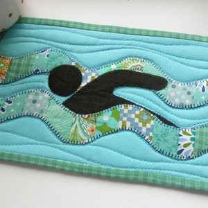 May include: A blue quilted coaster with a green border features a black silhouette of a swimmer in a wavy pattern of colorful fabric.