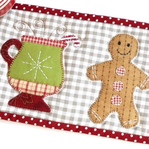 May include: A brown gingerbread man with red and white gingham buttons and a green mug with a red and white gingham trim and a candy cane on top. The mug and gingerbread man are on a grey and white gingham background with a red and white polka dot border.