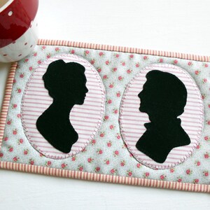 May include: A pair of fabric coasters with a pink and white striped background and a floral pattern. Each coaster features a black silhouette of a person's head in an oval frame.
