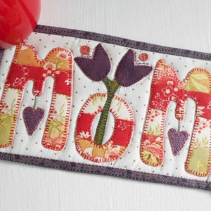 May include: A handmade quilted coaster with the word "MOM" spelled out in colorful fabric letters. The letters are surrounded by a purple border and a white background with small white dots. A purple flower with two leaves is centered between the letters.