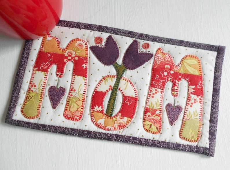 May include: A handmade quilted coaster with the word "MOM" spelled out in colorful fabric letters. The letters are surrounded by a purple border and a white background with small white dots. A purple flower with two leaves is centered between the letters.