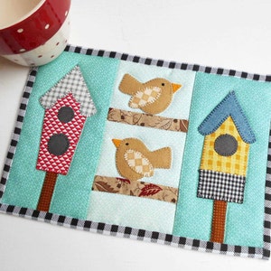 May include: A turquoise blue quilted coaster with a black and white checkered border. The coaster features three birdhouses with red, yellow, and blue accents, and two birds with brown and white checkered patterns.