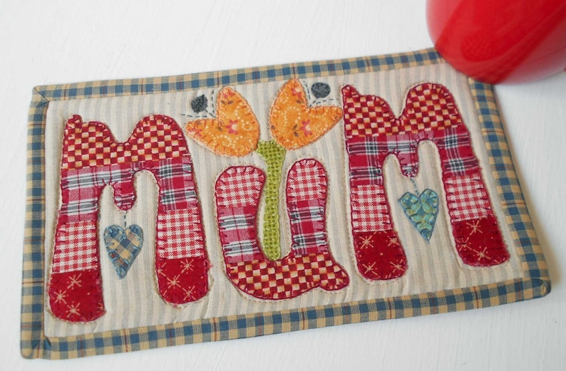 May include: A handmade quilted coaster with the word "MUM" in red, white, and blue patchwork fabric. The coaster features a green stem with two orange and yellow flowers.