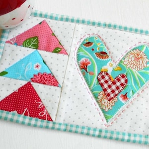 May include: A handmade quilted coaster with a heart design in the center. The heart is made of turquoise fabric with a red and white checkered heart in the center. The coaster is surrounded by a turquoise and white gingham border.