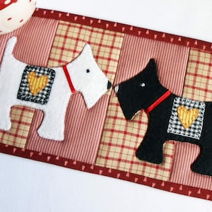 May include: A red, white, and black quilted coaster with two felt dogs, one white and one black, with red collars and yellow hearts on their chests. The coaster is made of patchwork fabric with a plaid pattern.