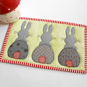 May include: A green and white quilted coaster with three gray bunny rabbits with red and white patterned bottoms. The coaster has a red and white striped border.