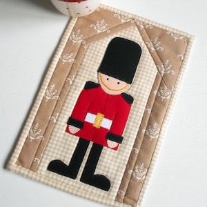 May include: A brown and beige quilted coaster with a red, white, and black felt appliqué of a British guard. The guard is wearing a black hat and a red coat with a white belt.