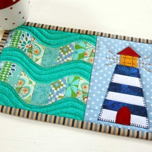 May include: A blue and white striped lighthouse with a red door and a yellow roof is appliqued on a blue fabric with white polka dots. The lighthouse is surrounded by a turquoise fabric with a wavy pattern. The fabric is bordered with a tan and navy blue striped trim.