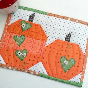 May include: A quilted orange and green pumpkin-shaped coaster with a white background. The coaster has a brown stem and three green hearts on a vine.