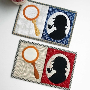 May include: Two quilted fabric coasters with a detective theme. The first coaster features a magnifying glass with the words "Thinking out loud" and "Wow!" inside the lens. The second coaster features a silhouette of a detective with a pipe and a hat. Both coasters have a checkered border.