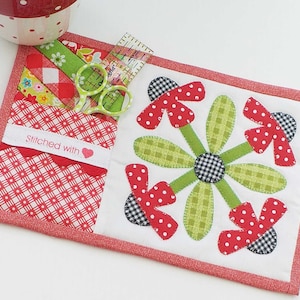 May include: A red and white quilted coaster with a pocket and a flower design. The pocket has a label that says "Stitched with" and a heart. The coaster is surrounded by a red border.