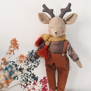 Könnte beinhalten: Eine Hirsch-Stoffpuppe mit geschlossenen Augen und rosa Wangen, bekleidet mit einem braunen Langarmshirt, einer rostfarbenen Latzhose mit zwei Knöpfen und einem gelben Strickschal mit roten Quasten.