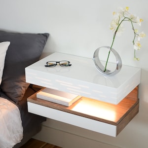 May include: A modern, floating nightstand with a glossy white top and a lower drawer with a wood grain finish. A pair of eyeglasses and a silver vase with a white flower are on top. A decorative pillow and a gray bedspread are in the background.