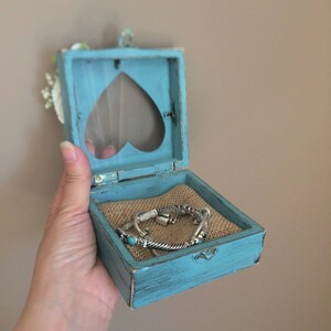Puede incluir: Una peque&ntilde;a caja de madera azul turquesa con un recorte en forma de coraz&oacute;n en la tapa. La caja est&aacute; abierta y contiene dos pulseras de plata con piedras turquesa.