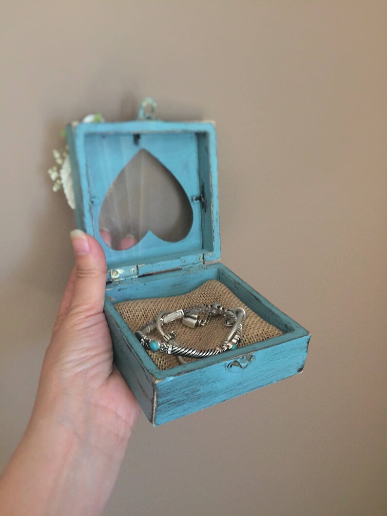 Puede incluir: Una peque&ntilde;a caja de madera azul turquesa con un recorte en forma de coraz&oacute;n en la tapa. La caja est&aacute; abierta y contiene dos pulseras de plata con piedras turquesa.