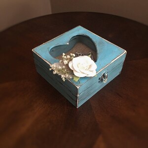 Puede incluir: Una peque&ntilde;a caja de madera azul con un recorte en forma de coraz&oacute;n. La caja est&aacute; decorada con pintura blanca y tiene un forro de arpillera. Una flor blanca y peque&ntilde;as flores blancas est&aacute;n dentro de la caja.