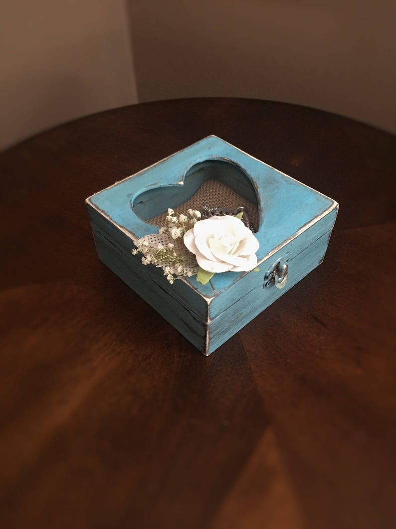 Puede incluir: Una peque&ntilde;a caja de madera azul con un recorte en forma de coraz&oacute;n. La caja est&aacute; decorada con pintura blanca y tiene un forro de arpillera. Una flor blanca y peque&ntilde;as flores blancas est&aacute;n dentro de la caja.