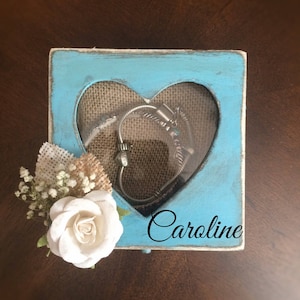 Puede incluir: Una caja de madera azul claro con un recorte en forma de coraz&oacute;n y una ventana transparente. La caja tiene un forro de arpillera y el nombre "Caroline" est&aacute; escrito en la parte delantera. Una rosa blanca y peque&ntilde;as flores blancas est&aacute;n unidas al lado de la caja.