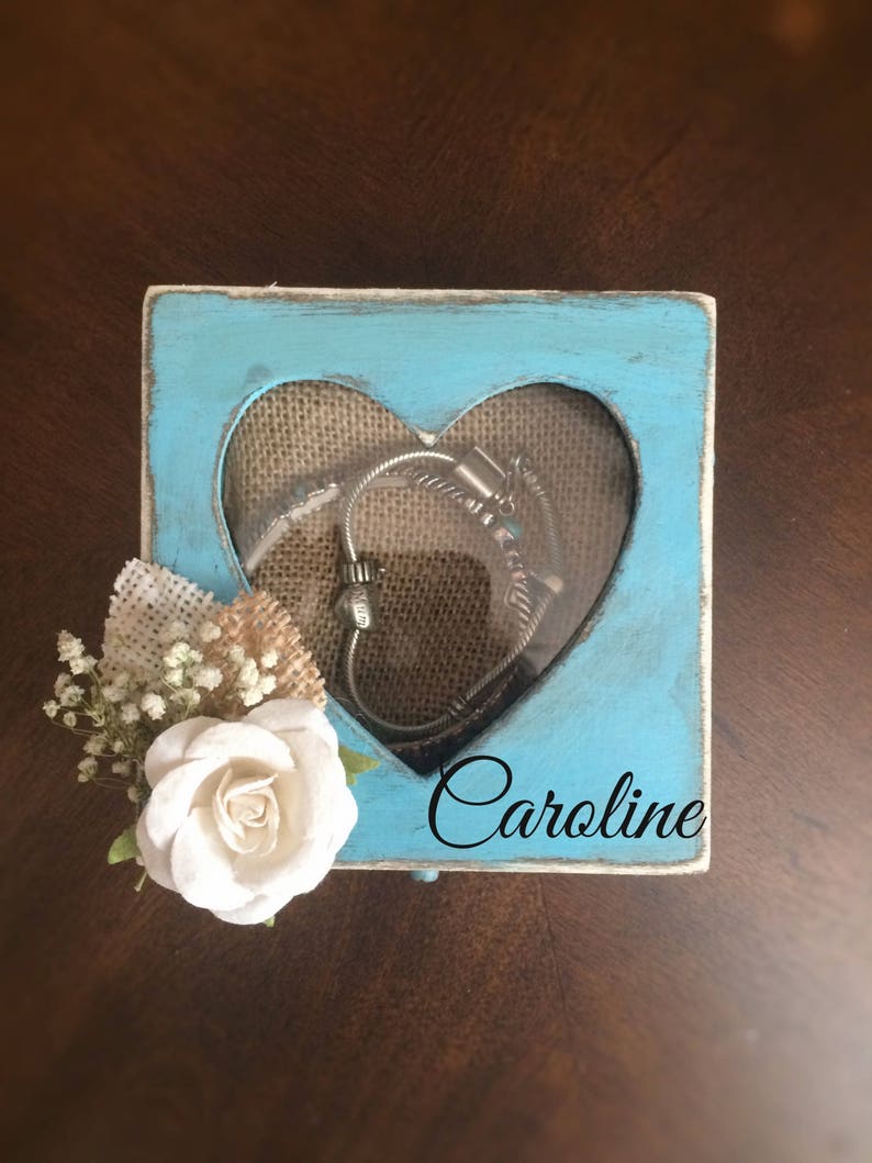 Puede incluir: Una caja de madera azul claro con un recorte en forma de coraz&oacute;n y una ventana transparente. La caja tiene un forro de arpillera y el nombre "Caroline" est&aacute; escrito en la parte delantera. Una rosa blanca y peque&ntilde;as flores blancas est&aacute;n unidas al lado de la caja.