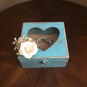 Puede incluir: Una peque&ntilde;a caja de madera azul turquesa con un recorte en forma de coraz&oacute;n en la tapa. La caja tiene un forro de arpillera y una peque&ntilde;a flor blanca en el lateral. La caja est&aacute; cerrada y tiene un cierre de metal.