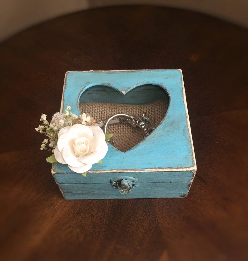Puede incluir: Una peque&ntilde;a caja de madera azul turquesa con un recorte en forma de coraz&oacute;n en la tapa. La caja tiene un forro de arpillera y una peque&ntilde;a flor blanca en el lateral. La caja est&aacute; cerrada y tiene un cierre de metal.