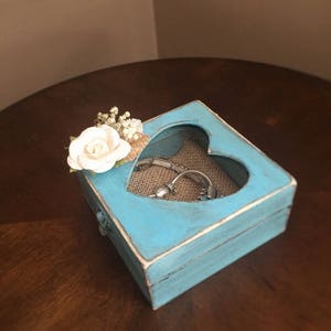 Puede incluir: Una caja de madera en forma de coraz&oacute;n de color azul claro con un acabado blanqueado. La caja tiene un coraz&oacute;n recortado en la parte delantera y un forro de arpillera en el interior. Una flor blanca y vegetaci&oacute;n se colocan en la parte superior de la caja.
