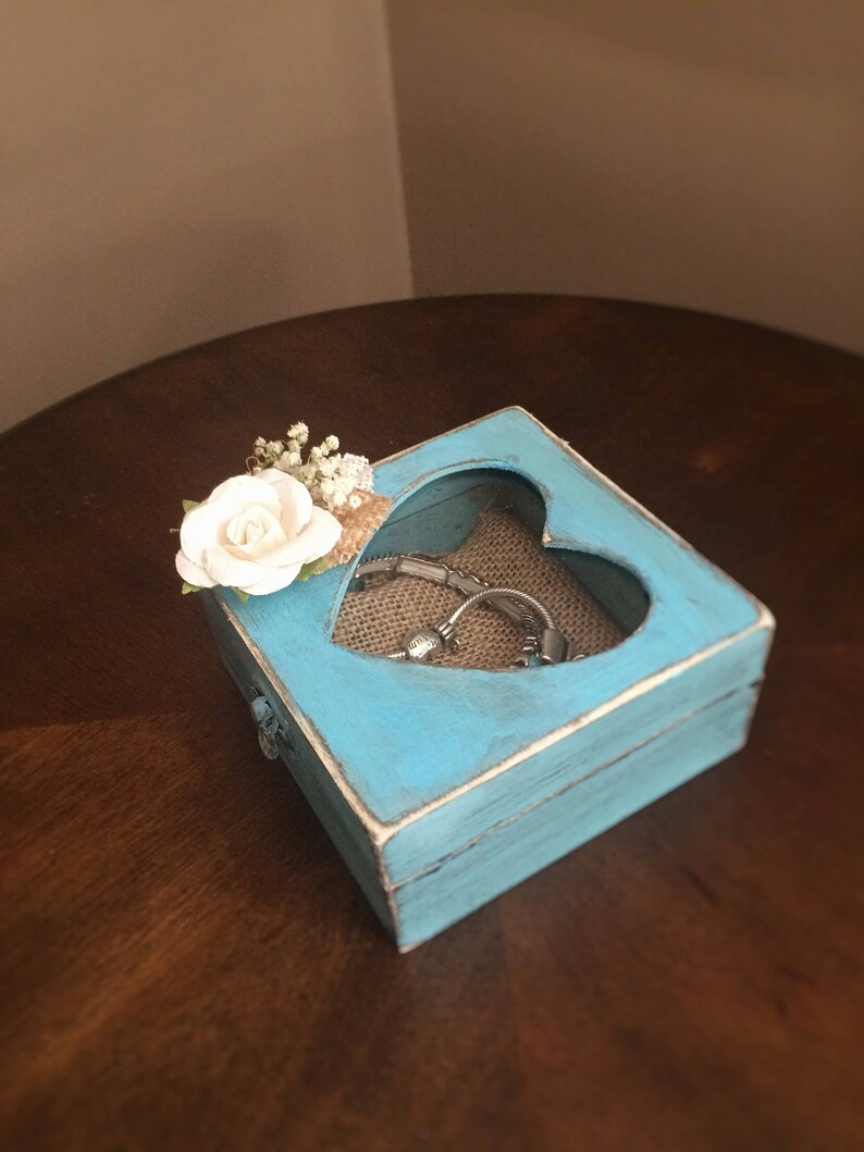 Puede incluir: Una caja de madera en forma de coraz&oacute;n de color azul claro con un acabado blanqueado. La caja tiene un coraz&oacute;n recortado en la parte delantera y un forro de arpillera en el interior. Una flor blanca y vegetaci&oacute;n se colocan en la parte superior de la caja.