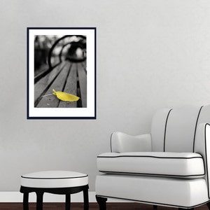 May include: A black and white photograph of a wooden bench with a single yellow leaf on it.