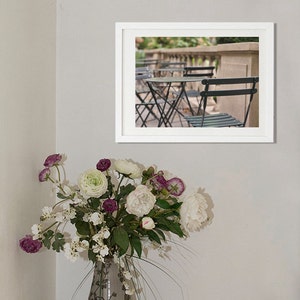 May include: A framed photograph of an outdoor cafe scene with black metal chairs and tables. The chairs and tables are arranged in a row, with a stone wall and greenery in the background.
