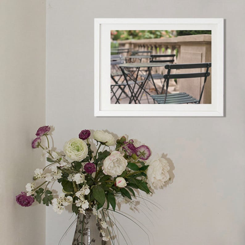 May include: A framed photograph of an outdoor cafe scene with black metal chairs and tables. The chairs and tables are arranged in a row, with a stone wall and greenery in the background.