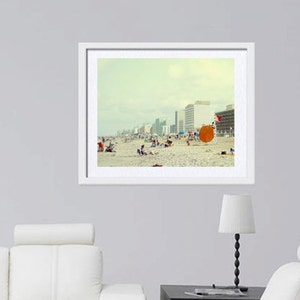 May include: A framed photograph of a beach scene with a group of people relaxing on the sand. The beach is lined with tall buildings and the sky is a pale blue. There is a large orange beach umbrella in the foreground.