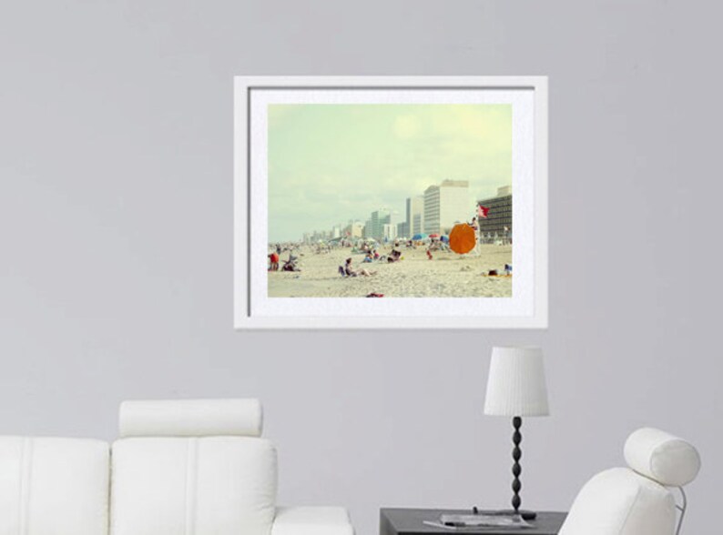May include: A framed photograph of a beach scene with a group of people relaxing on the sand. The beach is lined with tall buildings and the sky is a pale blue. There is a large orange beach umbrella in the foreground.